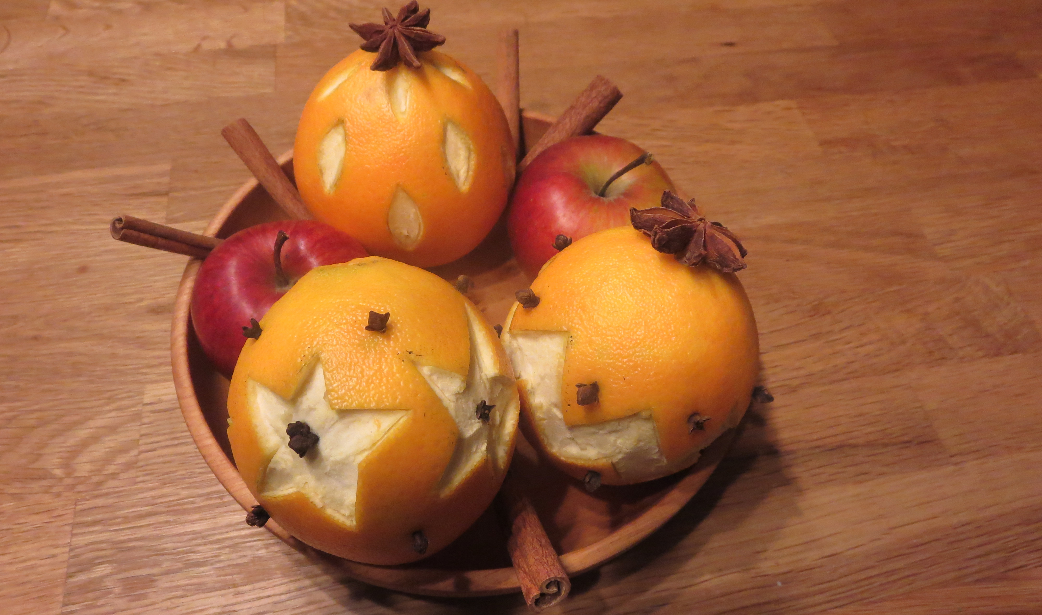 Drei geschnitzte Orangen sind mit zwei Äpfeln, Zimtstangen und Sternanis in einer Schale drapiert.