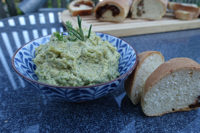 Eine Schüssel mit Zucchini-Aufstrich steht neben selbst gemachten Ciabatta auf dem blauen Tisch.