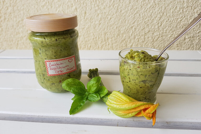 Neben dem verschlossenen Glas mit grünem Zucchini-Pesto steht ein offenes Glas davon, dekoriert mit einem Basilikumblatt und einer Zucchiniblüte.