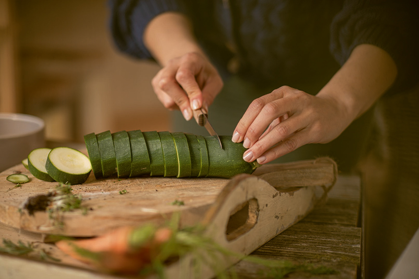 Auf einem Schneidebrett wird eine Zucchini mit einem Messer in Scheiben geschnitten.