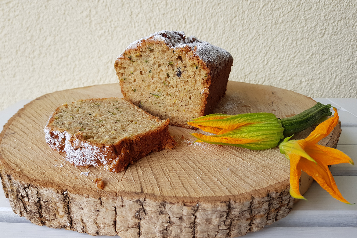 Der aufgeschnittene Zucchinikuchen liegt auf einer Holzscheibe und ist mit einer Zucchiniblüte garniert.