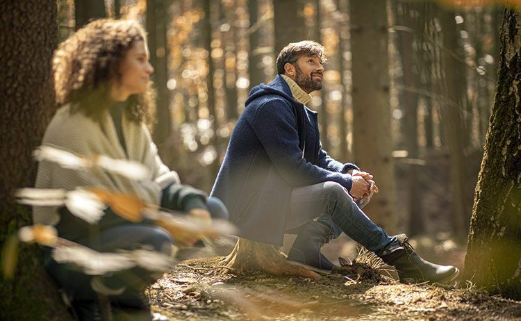 Ein Mann und eine Frau sitzen im Wald auf dem Boden und lassen die Natur auf sich wirken.
