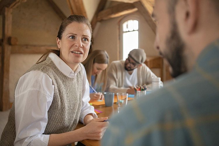 Eine Frau sitzt mit ihren Arbeitskolleginnen und -kollegen in einem Raum und diskutiert mit ihrem Gegenüber.