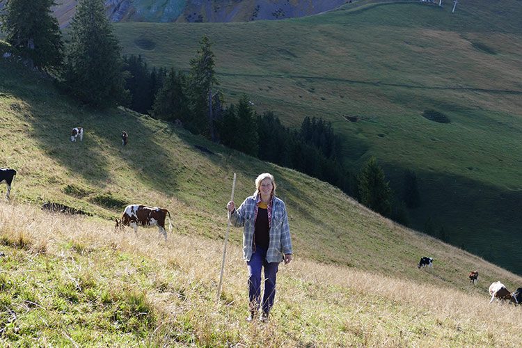 Katharina Afflerbach steht mit einem Stock in der Hand auf einer Kuhweide in den Bergen.