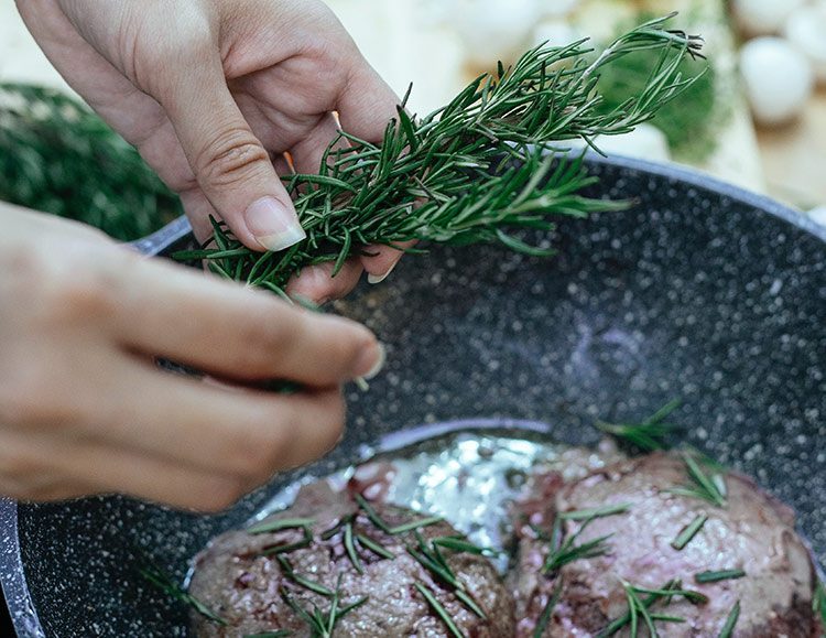 Eine Frau zupft Rosmarinblättchen von einem Zweig und gibt sie in eine Pfanne, in der Fleisch brät.