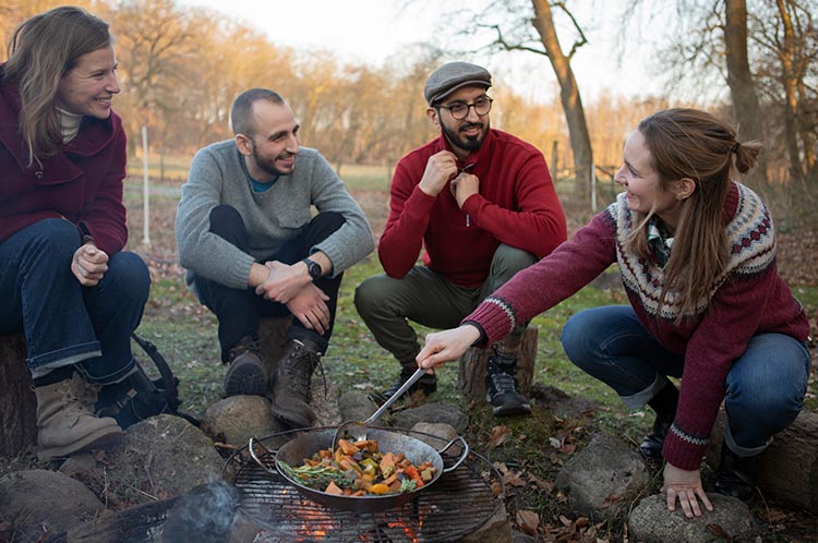 Eine Gruppe sitzt um ein Lagerfeuer herum und gart Gemüse in einer Pfanne auf dem Grillrost.