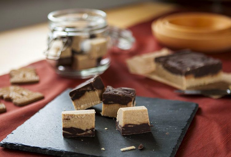 Der Spekulatius Fudge liegt auf einem Schieferbrettchen, im Hintergrund steht ein Glas mit dem selbst gemachtem Fudge.