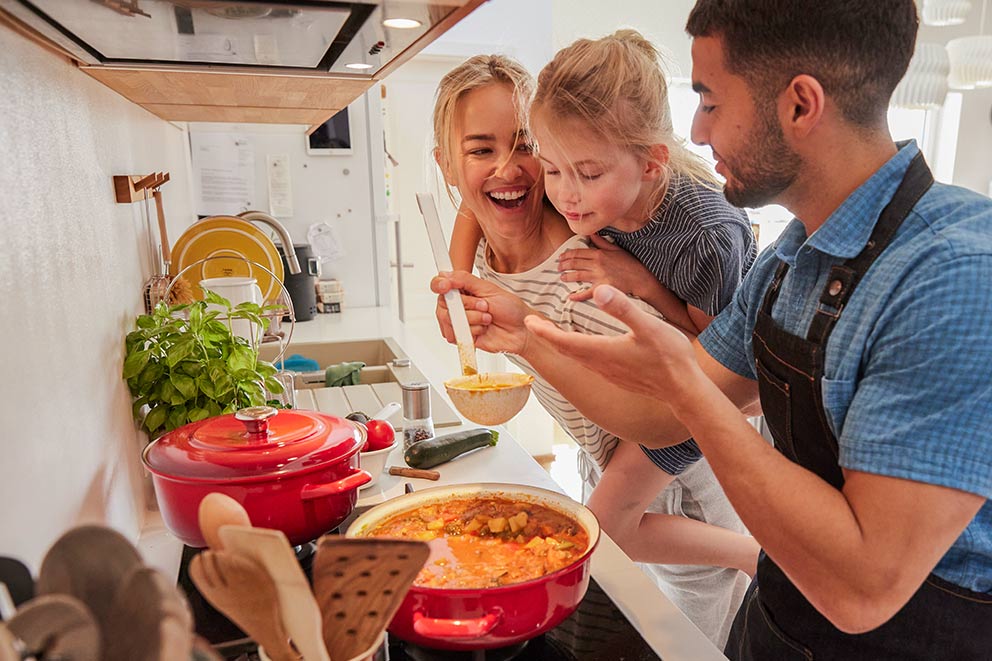 Das Kochen mit Kindern macht dieser Familie viel Spaß, sie stehen lachend vor dem Herd und probieren das Gericht.