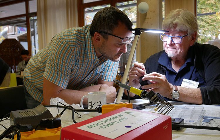 Zwei Männer beugen sich unter einer Lampe nah an einen kaputten Gegenstand, um ihn zu reparieren.