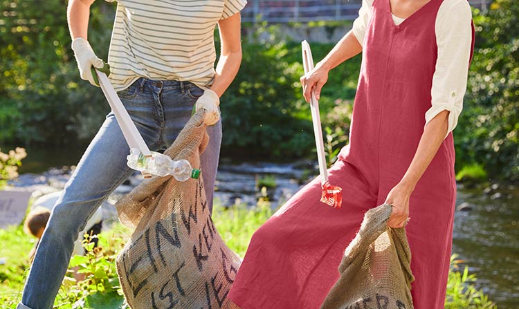 Zwei Frauen sammeln mit Beuteln und Zangen Müll in der Natur auf.