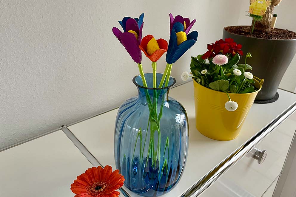 Die Blumen aus Eierkarton stehen in einer blauen Vase auf einer Kommode.