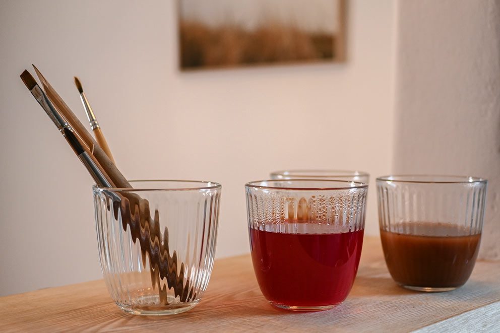 Selbst hergestellte Naturfarben stehen in Gläser gefüllt neben Pinseln auf dem Tisch.