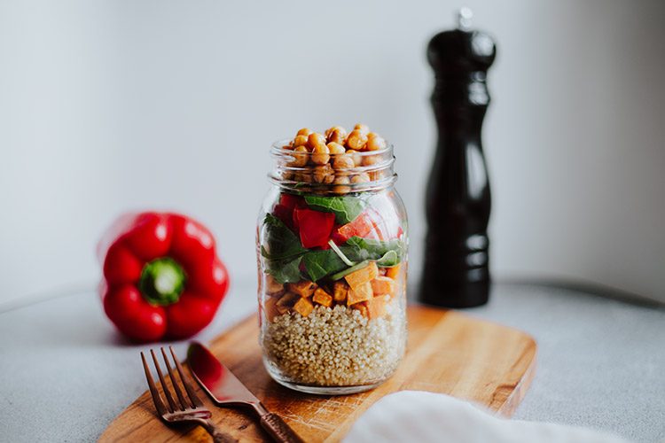 Ein Quinoa-Salat im Glas steht auf einem Holzschneidebrett, neben dem eine Paprika liegt.