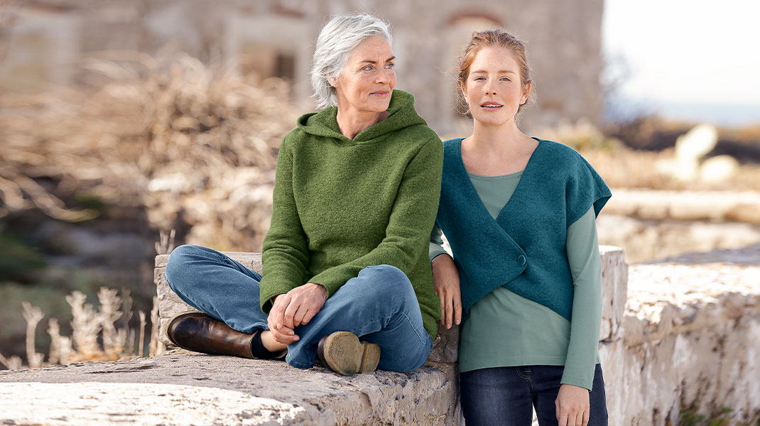 Eine ältere Frau sitzt auf einer alten Mauer und trägt einen Walk-Kapuzenpullover. Neben ihr lehnt eine jüngere Frau und  trägt eine Walk-Weste
