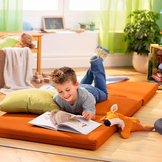 Junge liest liegend auf seiner orangen Matratze in einem nachhaltig eingerichteten Kinderzimmer