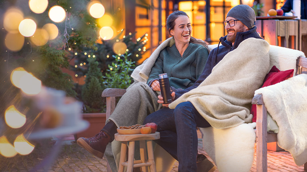 Eine Frau und ein Mann sitzen in Decken eingekuschelt draußen auf der Bank in weihnachtlicher Atmosphäre.